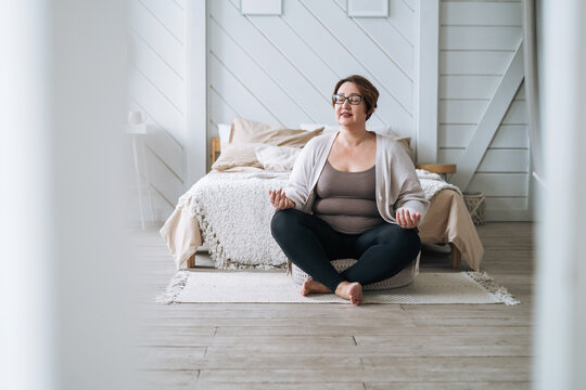 Middle aged plus size smiling brunette woman in home wear practicing yoga in bedroom