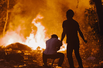 Two men are extinguishing a fire in a tropical forest, one of them is hosing down. The forest is red, all on fire, a lot of smoke.