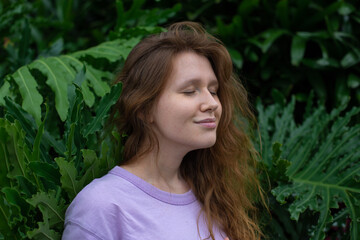 Naklejka premium Portrait of young woman standing on an ivy, fern wall or in greenery, breathing, smiling and looking away girl relaxing outdoors at summer day.Love for nature, eco life,green leaves, green park