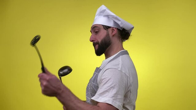 Cheerful male cook holding kitchen utensils, having fun isolated on yellow background. Positive man chef enjoying cooking, playing by implement tools like drumsticks indoors. Culinary concept