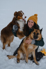 Girl plays with dogs outside in winter. Young pretty Caucasian woman sitting in snow and cuddling with two dogs - German and Australian Shepherds. Concept of warm relationship between owner and pets.