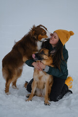 Concept of warm relationship between owner and pets. Girl kisses dog outside in winter. Young pretty Caucasian woman sitting in snow and cuddling with two dogs - German and Australian Shepherds.