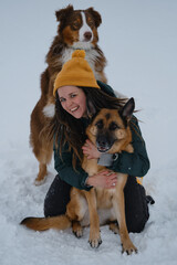 Girl plays with dogs outside in winter. Young pretty Caucasian woman sitting in snow and cuddling with two dogs - German and Australian Shepherds. Concept of warm relationship between owner and pets.