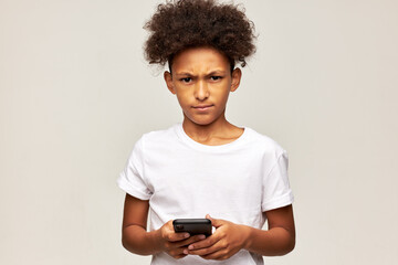 Angry african american kid boy in white t-shirt upset with him losing online game on his smartphone standing against gray studio background, looking at camera with annoyed facial expression