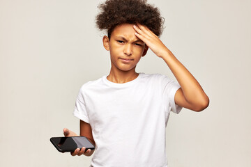Fototapeta African american kid boy suffering headache touching forehead after playing mobile games too much, standing against gray studio background with smartphone in hands, dressed in white mockup t-shirt obraz