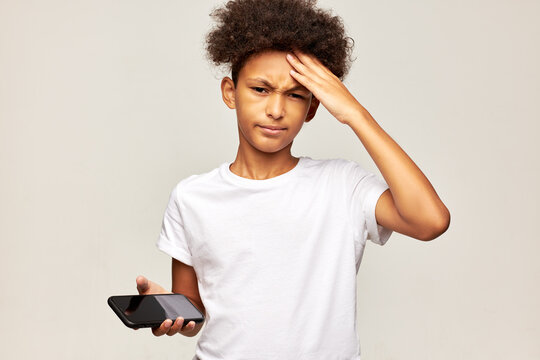 African american kid boy suffering headache touching forehead after playing mobile games too much, standing against gray studio background with smartphone in hands, dressed in white mockup t-shirt