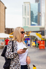 Fototapeta premium Amidst the modern architece, lively harbor, luxurious boats, towering skyscrapers, and sunny sky with clouds, a blonde tourist girl wanders around the Dubai resort area, taking selfies along the way.