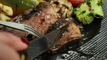 The knife cuts off piece of steak against the background of grilled vegetables