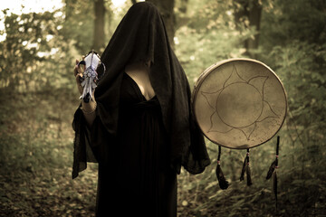 Young witch in twilight forest holding shamanic drum and wolf skull. A mysterious pagan priestess with covered face. Mystic Halloween witchy atmosphere. Occulture, magic, spiritual, witchcraft.