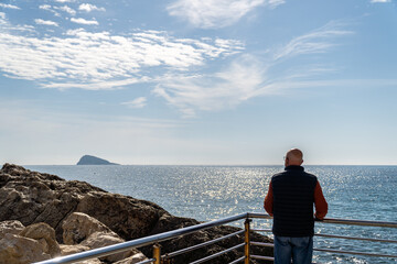  A mature man with his back turned, leaning on a metal railing watching the sea with an island in the background, in Benidorm, Alicante (Spain)
