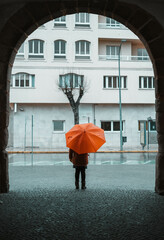 Girl with an orange umbrella standing still under an arch watching how it rains in the city