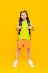 A child with a school backpack. A beautiful little girl in full height, in shorts and a T-shirt. Education during the summer holidays. Preparatory courses for children. Yellow isolated background.