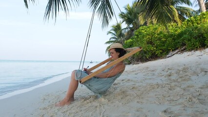 Young woman in straw hat making notes while resting in swings on tropical beach