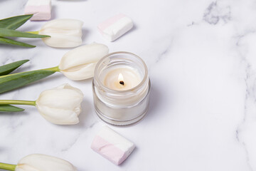 Fototapeta na wymiar Soy wax candle in glass on marble background. White tulips and marshmallows