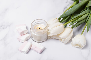 Obraz na płótnie Canvas Soy wax candle in glass on marble background. White tulips and marshmallows. Scented candle