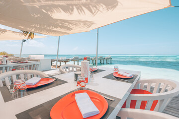 Obraz premium Relaxing outdoor restaurant at the beach. Table setting at tropical beach restaurant. Elegant white wooden tables, chairs under beautiful soft sunset sky, sea view. Luxury hotel or resort restaurant