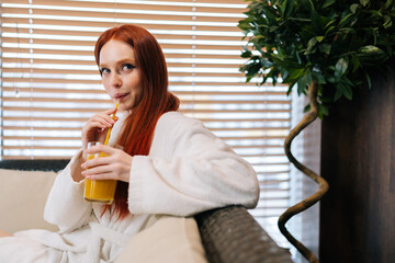 Close-up portrait of pretty young woman in white bathrobe drinking cocktail from straw sitting on sofa by window at spa resort. Charming redhead female relaxing after body treatment at beauty salon.