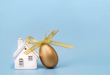 Happy Easter concept. One golden egg with a ribbon bow and white ceramic house on blue. Copy space
