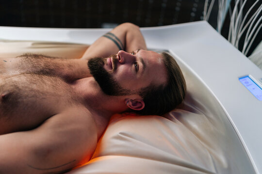 Close-up side view of bearded young man lying relaxing on electric massage bed and enjoying spa treatment at luxury spa salon. Muscular male having rest enjoying wellness weekend, thinking looking up.