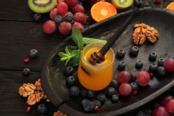 Natural golden honey in a glass bowl on a wooden table. The honey is decorated with berries, fruits, mint greens and nuts.