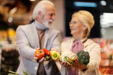 Close up of senior couple showing fresh vegetables at the camera while standing at the hypermarket.