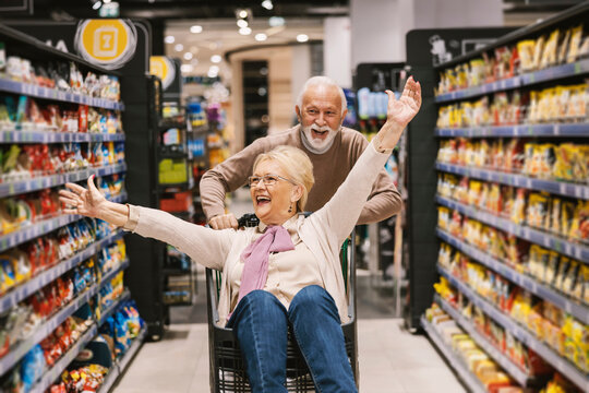 A playful senior couple in love is riding shopping cart at the supermarket and having fun.