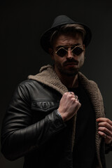 sexy fashion man with hat and glasses adjusting wool cardigan and leather jacket