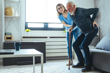 Smiling nurse assisting senior man to get up from bed. Caring nurse supporting patient while getting up from bed and move. Helping elderly disabled man standing up in his living room.