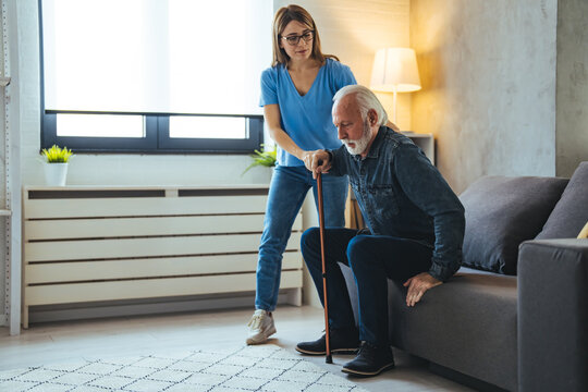 Smiling senior man with female healthcare worker. Home carer supporting old man to stand up from the chair at care home.. Female caregiver helping senior man get up from couch in living room - Indoors