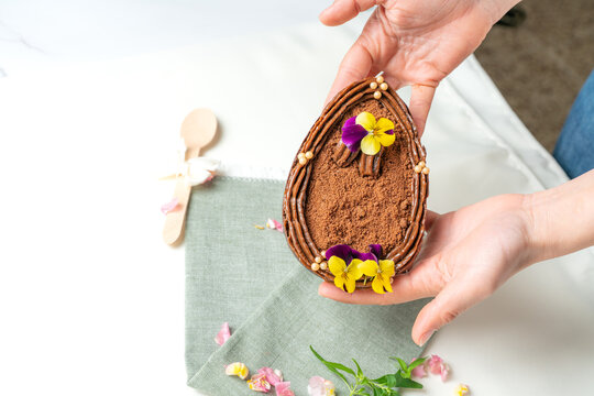 Hands holding a presentation of an Easter egg from a pastry stuffed with chocolate and caramel, and decorated with showy yellow flowers...