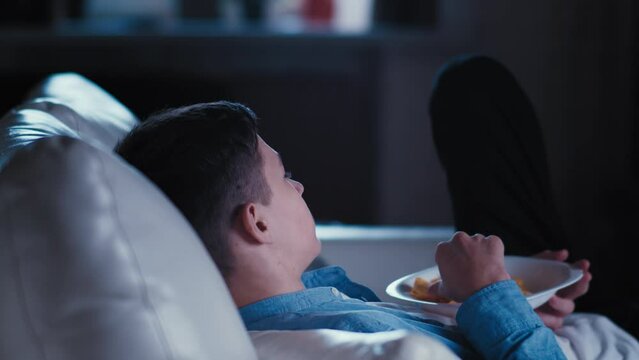 Teenage Boy Relaxes at Home: Watching TV, Snacking on Potato Chips, and Channel Surfing