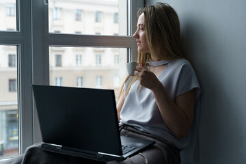 A young woman sits on a windowsill, drinks coffee, works at a laptop, looks out the window.