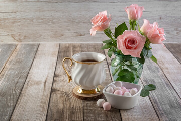 Pink roses in a glass vase, a white cup and round candies on a wooden table. Postcard. Copy space. Photo