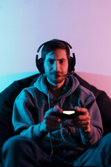 Young smiling male gamer wearing headset with mic and using controller while sitting on pouf...