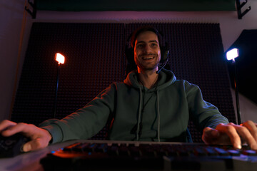Fototapeta na wymiar Low angle of young cheerful cybersport gamer in headset using keyboard and mouse while playing video game on computer in dark room illuminated by neon lights