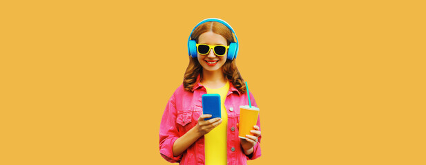 Portrait of stylish modern happy smiling young woman in headphones listening to music with smartphone wearing pink jacket on orange background