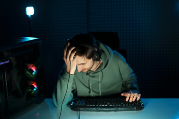Young male cybersport gamer in headset with microphone using keyboard while playing video game in dark room illuminated by neon light looking at camera