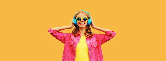 Portrait of happy smiling young woman in headphones listening to music wearing pink jacket on orange background