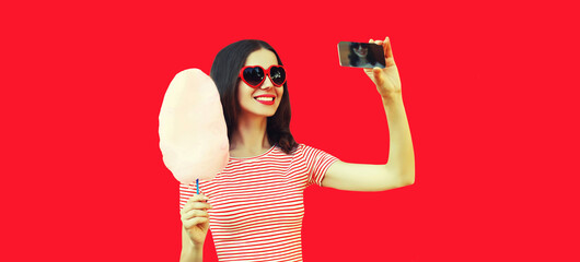 Portrait of happy smiling young woman with cotton candy taking selfie with smartphone wearing red heart shaped sunglasses on background