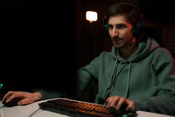 Concentrated male cybersport gamer wearing headset and playing video game having battle in dark room illuminated by neon lights