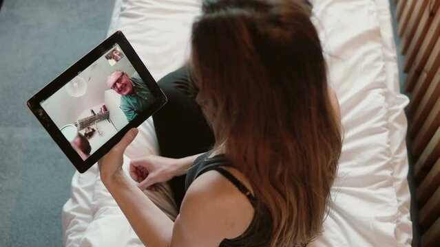 Young woman having a video call over a tablet. Attractive girl sitting at bed and talking with father, video conference.