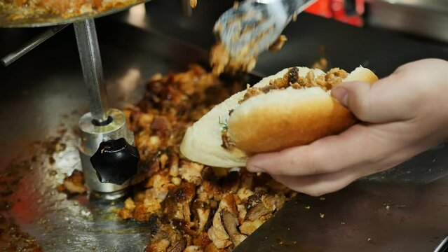 The hands of a fast food chef holding culinary tongs put pieces of fried juicy meat into a cut bun for making doner kebab with vegetables and sauce. Muffins and fatty meats are harmful