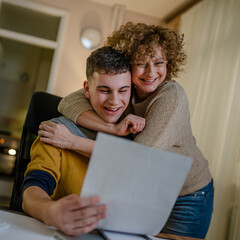 Young man and his mother woman hold paper read good news in letter sit at desk at home beautiful...