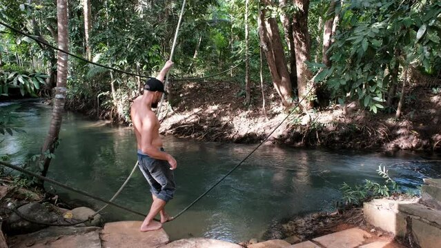 Tripod shot of a tourist (middle aged man) walking a tightrope over crystal clear creek in the jungle on sunny day. Krabi Province, Thailand.