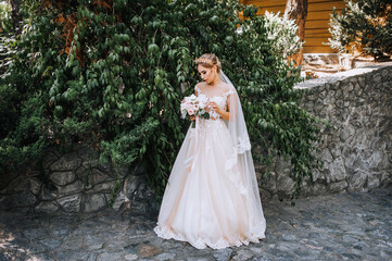 A beautiful blonde model bride in a white lace dress with a crown, a diadem on her head stands in a park in nature with a bouquet in her hands. Wedding photography, portrait.