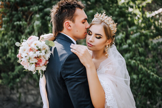Stylish groom in a blue suit and a beautiful blonde princess model bride with a crown with a bouquet hug together while standing in the park outdoors. Wedding photography of newlyweds, portrait.