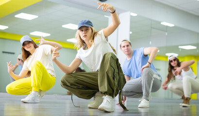 Expressive teen girl and three teenagers in caps sitting in breakdance pose in dance hall