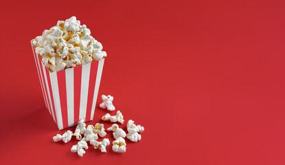 Red white striped carton bucket with tasty cheese popcorn, isolated on red background. Box with scattering of popcorn grains. Fast food, movies, cinema and entertainment concept.