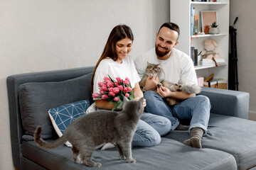 A couple in love is sitting with cats and pink tulips on the sofa at home.