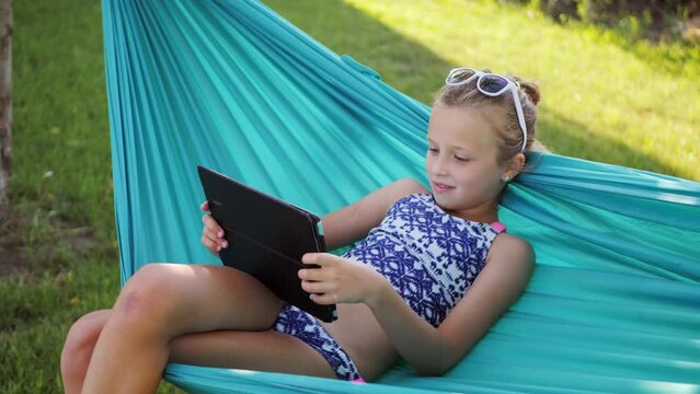 Side view of a young woman smiling looking at smartphone screen is in her hands, while lying in the orange hammock.Film effect, blurred foreground and background.Girl using gadget